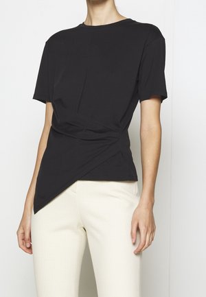 Schwarzes T-Shirt mit kurzen Ärmeln, asymmetrischem Design und verdrehtem Vorderteil, kombiniert mit cremefarbener strukturierter Hose.