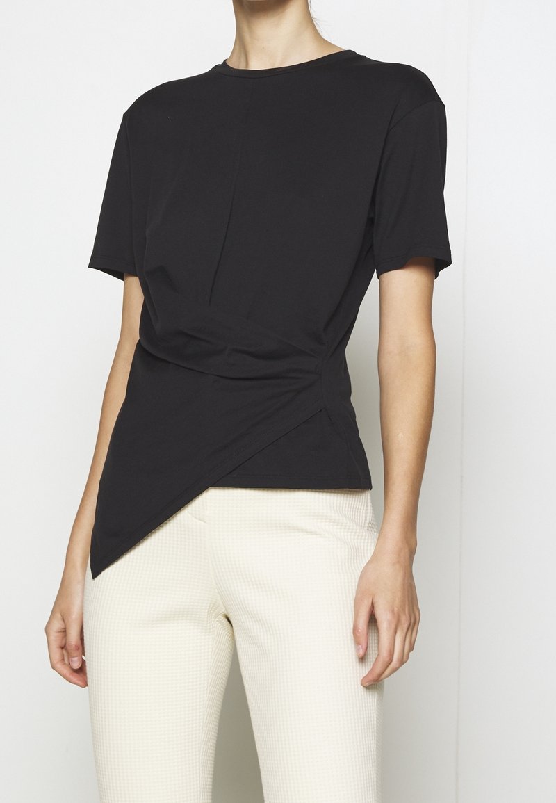 T-shirt noir à manches courtes avec un design asymétrique et un détail avant torsadé, associé à un pantalon crème texturé.