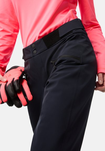 Marineblaue Skihosen verfügen über einen Klettverschluss und einen Knopfverschluss am Bund, seitliche Reißverschlusstaschen und werden mit leuchtend pinken Handschuhen für einen Kontrast kombiniert.