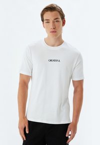 Hvid bomuld t-shirt med korte ærmer, rund halsudskæring og sort broderet tekst "GRATEFUL" på fronten. Glat tekstur, afslappet pasform.