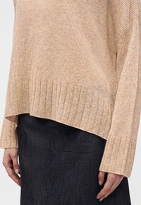 Personne portant un pull en maille côtelée beige à manches longues et une jupe en jean noire, montrée du torse au milieu de la cuisse.