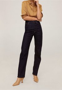 Jean bleu foncé taille haute avec une coupe droite et des coutures apparentes, associé à une chemise moutarde à manches courtes boutonnée et des talons beiges.