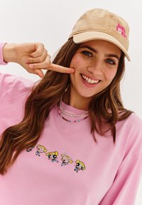 Růžový svetr má na sobě grafiku animovaných postaviček, béžová baseballová čepice pak zobrazuje růžový text. Přítomny jsou také vrstvené perlové náhrdelníky.