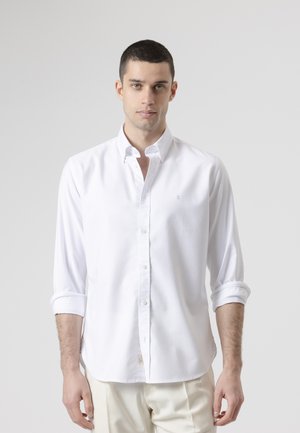 Joven con cabello corto y oscuro, vestido con una camisa blanca de botones y mangas remangadas, y pantalones claros, posando frente a un fondo liso.