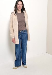 Giacca in pile beige con cappuccio, top marrone e jeans blu, caratterizzata da una vestibilità leggermente ampia e sneakers bianche con suola beige.