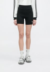 Person wearing black high-waisted shorts, white socks with black logo, gray slip-on athletic shoes, and a white long-sleeve top with black stripes.