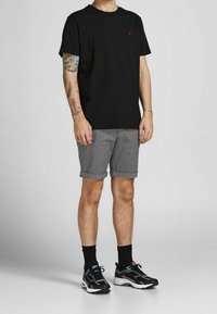 Mężczyzna w czarnym podkoszulku, szarych szortach z podwiniętymi nogawkami, czarnych skarpetkach i czarnych butach sportowych stoi z widocznymi tatuażami na lewej ręce na jednolitym tle.