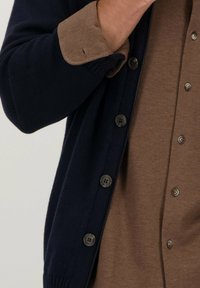 Marine gebreide cardigan met een bruine overhemd eronder. De cardigan heeft een knoopsluiting en ribbels aan de mouwen, het overhemd heeft een knoopsluiting.