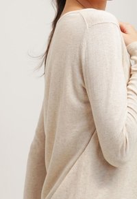 Pull beige à manches longues en maille, avec un col arrondi, des poignets côtelés et une texture lisse, présenté de profil avec le tissu tombant naturellement.