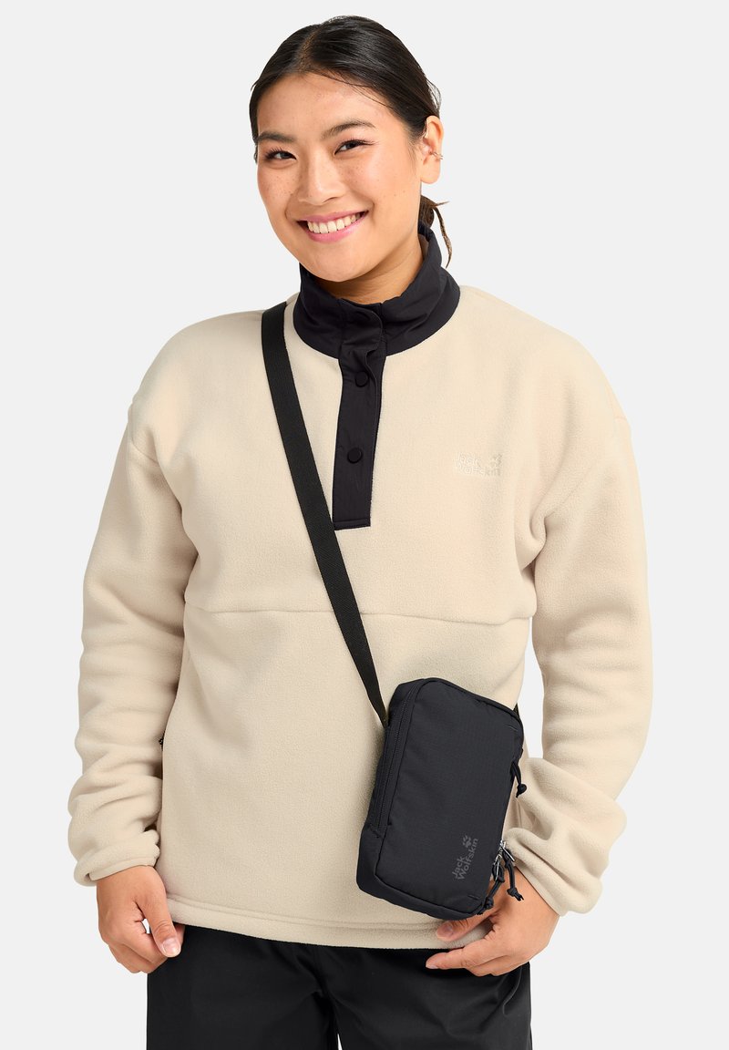 Personne souriante portant un pull en polaire beige avec un col noir et un sac bandoulière noir, se tenant devant un fond blanc uni.