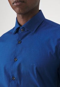 Camicia blu con bottoni, realizzata in tessuto testurizzato, colletto elegante e bottoni scuri; un logo discreto è ricamato sul petto.