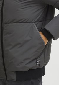 Dark gray padded jacket with a zip closure, black ribbed cuffs, and a side pocket with a logo tag. Smooth exterior texture.