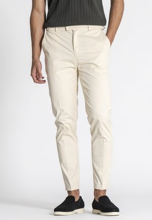 Mand iført beige slim fit-bukser med lommer, sorte slip-on sko og en mørk, ærmeløs ribstrikket top, stående mod en hvid baggrund.