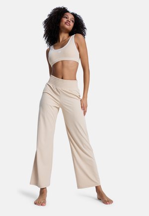 Kobieta o kręconych włosach, ubrana w beżowy prążkowany krótki top i szerokie spodnie, stojąca boso na białym tle.