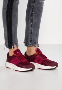 Burgundowe buty sportowe z siateczkową i syntetyczną cholewką, z gwiazdkowymi detalami, okrągłym noskiem i białą podeszwą. Noszone z postrzępionymi szarymi dżinsami.