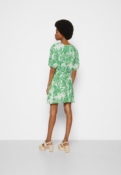 Vestido floral verde com mangas curtas bufantes, cintura ajustada e barra reta. Usado com sandálias de salto bloco bege.