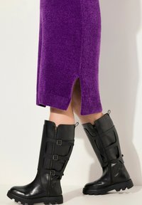 Stivali neri alti fino al ginocchio con una finitura liscia e lucida, dettagli con fibbie e cerniere, abbinati a un vestito di maglia testurizzata viola con spacchi laterali.