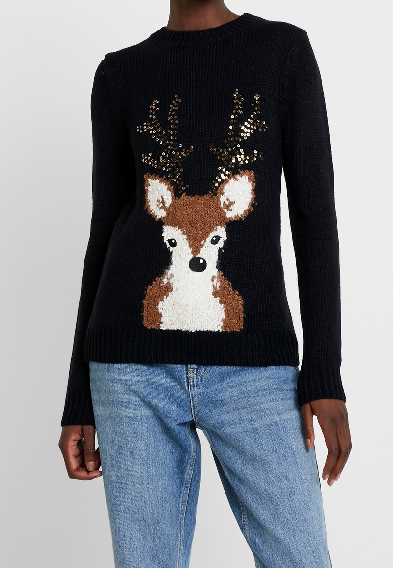 Schwarzer Strickpullover mit einem Hirschdesign, das strukturiertes Fell, pailettenbesetzte Geweih und gerippte Bündchen sowie Saum aufweist. Kombiniert mit blauen Jeans.