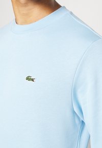 Felpa crewneck azzurro chiaro in cotone, con un piccolo logo di coccodrillo ricamato in verde sul petto. Texture liscia e design aderente.