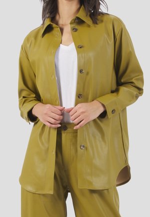 Vrouw draagt een openknopbaar, olijfgroen faux lederen overhemd en bijpassende broek over een wit T-shirt met een V-hals, handen dichtbij de knopen van het overhemd.