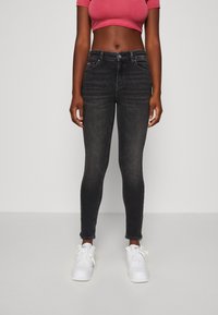 Jeans negros ajustados con cintura de media altura, desgaste sutil, cinco bolsillos y cremalleras en los tobillos, combinados con una blusa corta rosa y zapatillas blancas.