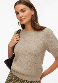 Pull beige en maille à manches courtes avec un motif texturé, encolure ronde et ourlet côtelé, accompagné d'un sac à main foncé.