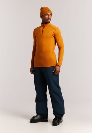Man draagt een oranje muts en een top met lange mouwen, donkerblauwe broek en zwarte skischoenen, staand voor een effen achtergrond.