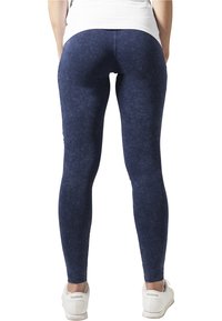 En person som står med ryggen vänd, iklädd mörkblå leggings, vita sneakers och en vit topp, mot en vit bakgrund.