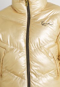 Złota kurtka puchowa z błyszczącą, odblaskową powierzchnią, czarnym zamkiem błyskawicznym i haftowanym logo na piersi. Miękka, pikowana tekstura z kołnierzem typu stójka.