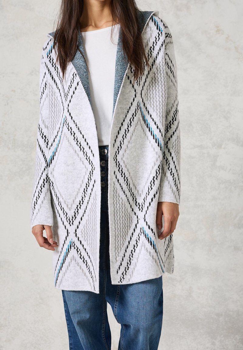 Cardigan en maille blanc avec motifs en losange bleus et noirs. Comprend une capuche, une ouverture sur le devant et des manches longues. Tissu texturé.