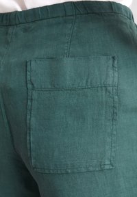 Pantalons en lin turquoise avec une poche arrière. Le tissu a une texture douce et un aspect légèrement froissé, agrémenté de bords surpiqués.