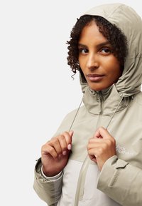 Lichtbeige waterdichte jas met een capuchon, voorzien van een rits aan de voorkant en koordjes, met een gestructureerde stof en een logo op de borst.