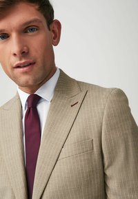 Beige gestreifter Sakk mit einer einzelnen Brusttasche, kombiniert mit einem weißen Hemd und einer dunkelburgunderfarbenen Krawatte. Glatte Textur.