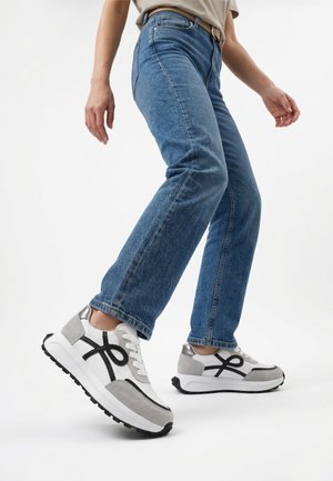 Person wearing blue jeans and white sneakers with black and gray details, walking against a plain white background.