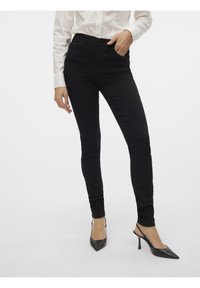 Femme portant un pantalon noir ajusté et des escarpins noirs pointus, debout devant un fond clair uni.