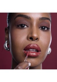 Läppglans i en djup röd nyans appliceras med en borste, vilket framhäver en jämn, glansig textur. Bakgrunden är en enfärgad mörk burgundy.