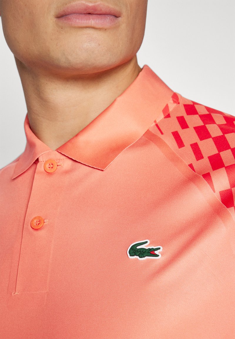 Kurzarm-Polo in hellem Koralle, mit glattem Material, einer orangefarbenen Knopfleiste und einem grünen Krokodil-Logo auf der Brust. Rote gemusterte Akzente.