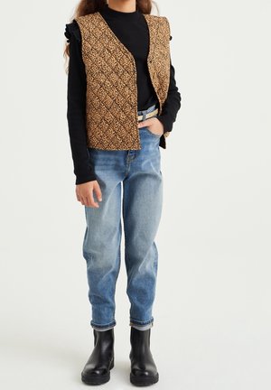 Beige gesteppte Weste mit Rautenmuster, schwarzes Langarmshirt, hellblaue Jeans und schwarze Stiefeletten.