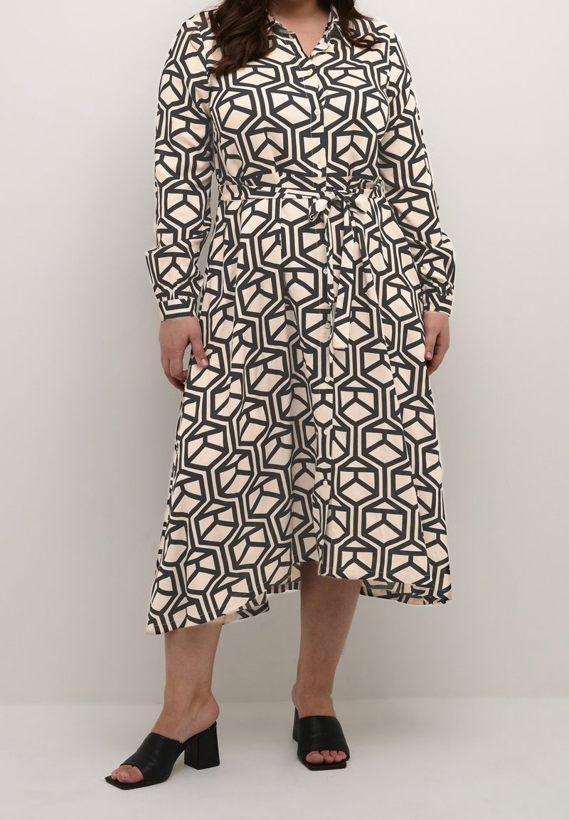 Femme portant une robe midi à manches longues à boutons, avec un motif géométrique noir et blanc et une ceinture à la taille, associée à des escarpins noirs à bout ouvert.