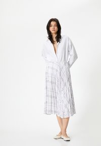 Chemise blanche à manches longues avec boutons, assortie à une jupe plissée de longueur genoux avec un motif à carreaux violet clair et des chaussures plates neutres.