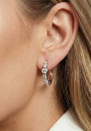 Nærbillede af en kvindes øre, der bærer en sølvørering i en ringform pyntet med syv små, runde, klare ædelsten.