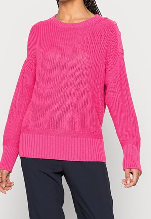 Nainen, joka pukeutunut kirkkaanpunaiseen neulottuun villapaitaan, jossa on napit yksityiskohtina yhdellä olkapäällä, ja mustiin housuihin, yksinkertaisen taustan edessä.