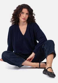Pull en V bleu marine en tissu doux, associé à un pantalon large en denim foncé. Chaussures plates noires avec bride à la cheville, design minimaliste.