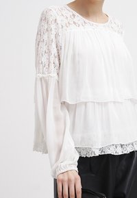 Blouse blanche à plusieurs couches avec des détails en dentelle le long de l'encolure et de l'ourlet ; manches longues transparentes ; tissu doux et léger avec des accents texturés.