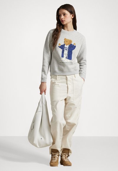 Grauer Sweatshirt mit einem Bärenmotiv in einem blauen Mantel, kombiniert mit cremefarbigen weit geschnittenen Hosen und braunen Schnürstiefeln, die eine weiße Tasche halten.
