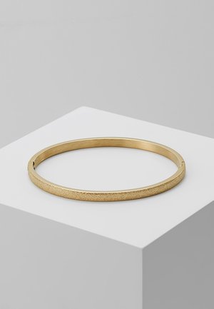 Bracelet jonc texturé doré avec fermoir, exposé sur un cube blanc, sur un fond gris uni.