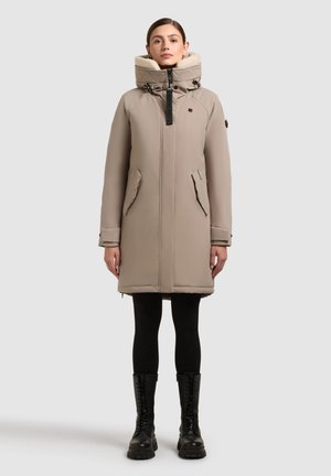 Beige long puffer coat with a high collar, zip front, side pockets, and black hardware. Styled with black leggings and lace-up black boots.