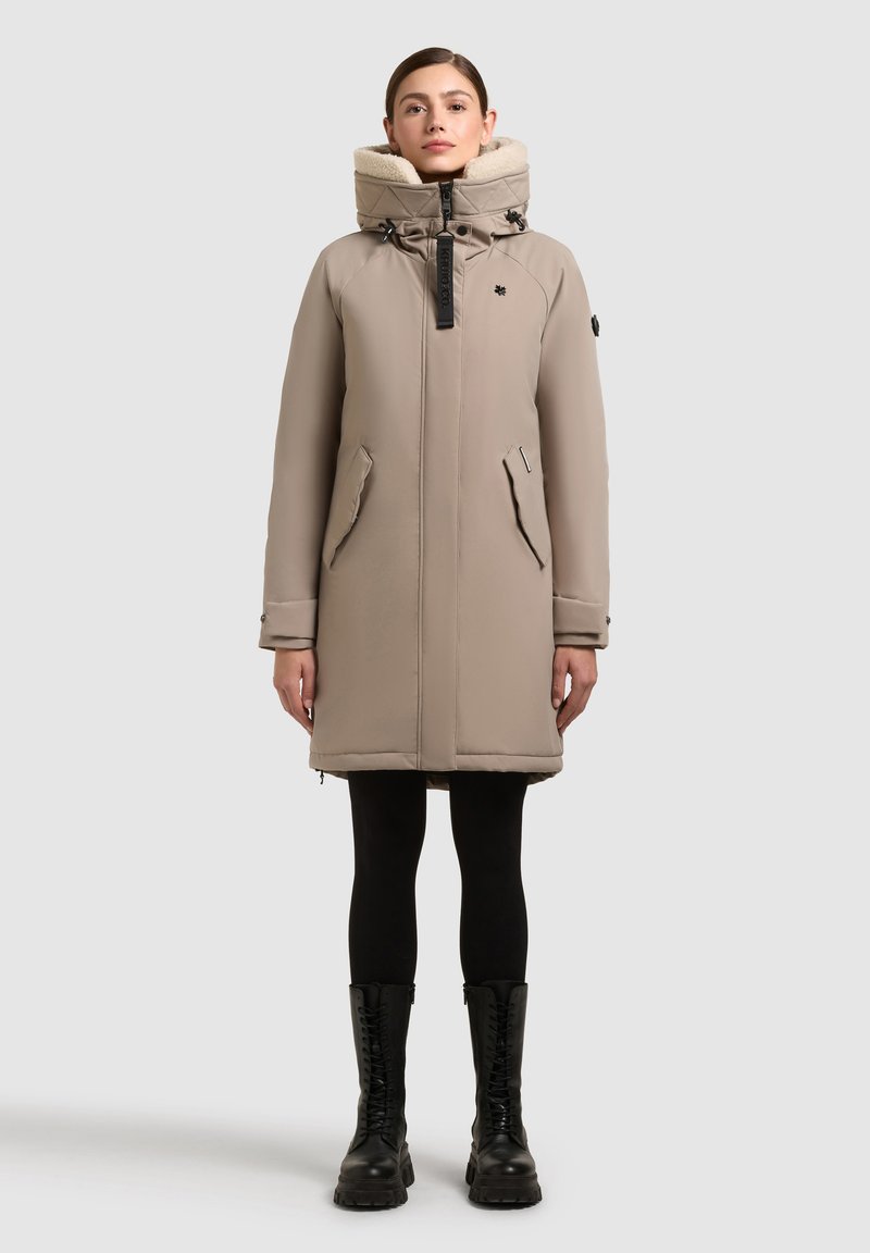 Cappotto lungo beige invernale con colletto alto, frontale con zip, tasche laterali e accessori neri. Abbinato a leggings neri e stivali neri con lacci.