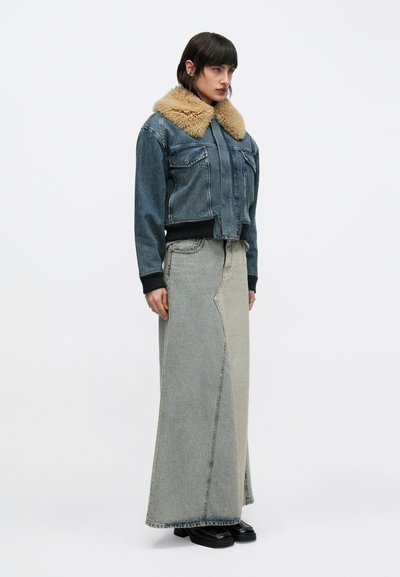 Moteris dėvi džinsinę striukę su rudos spalvos kailio apykakle, ilgą dviejų atspalvių džinsinę sijoną ir juodus platforminius batus, stovinti priešais paprastą foną.