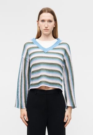 Kobieta ubrana w luźny, pasiasty, pastelowy sweter z dzianiny z dekoltem w serek, nałożony na białą koszulę, zestawiony z czarnymi spodniami z wysokim stanem.